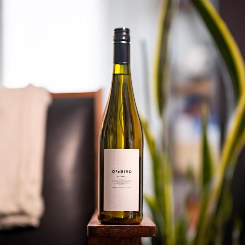 Bottle of Oddbird Presence Non-Alcoholic White Wine standing upright on a wooden chair with blurred background
