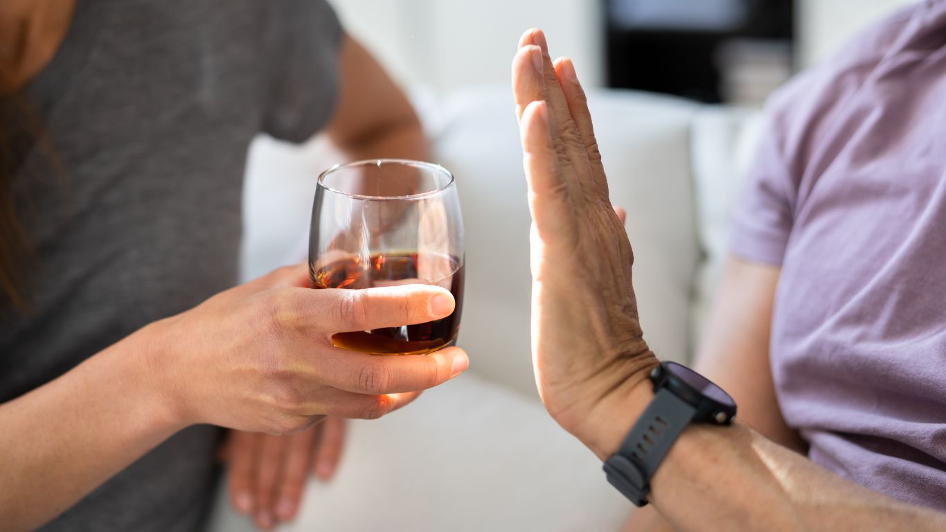 Person refusing an alcoholic drink by holding up their hand while another offers a glass of dark liquid