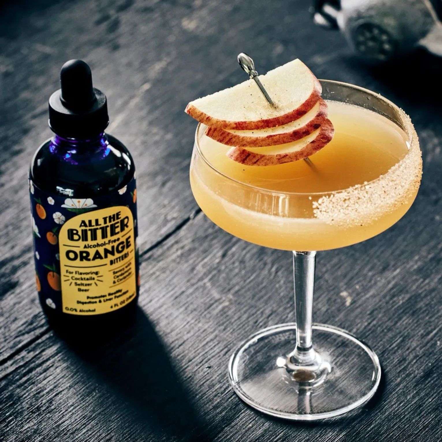 All The Bitter Orange Bitters bottle with golden non-alcoholic cocktail garnished with apple slices
