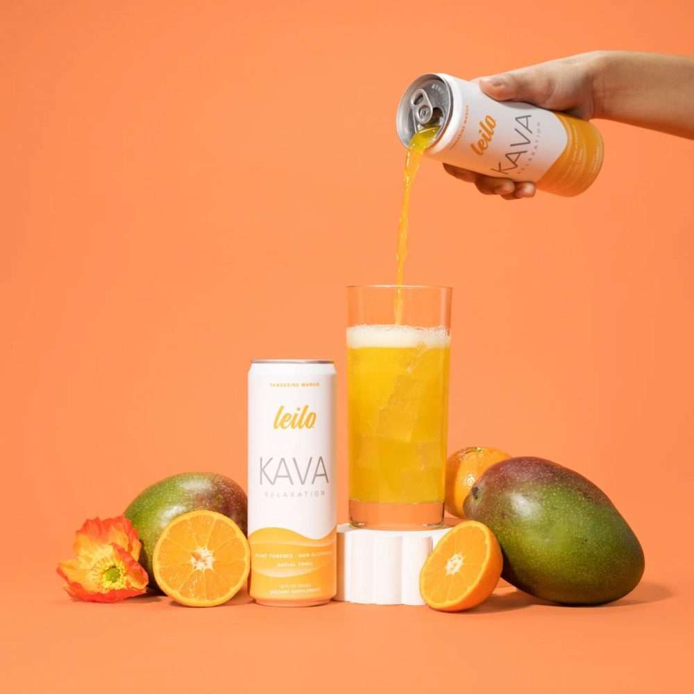 Hand pouring Leilo Tangerine Mango Kava drink into a glass with ice, mango, and citrus fruit.