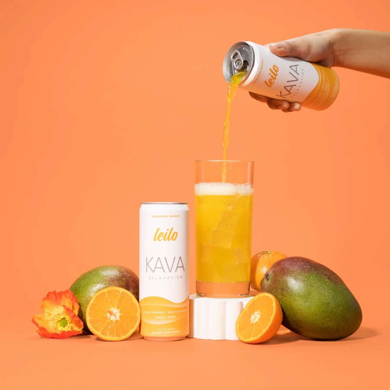 Hand pouring Leilo Tangerine Mango Kava drink into a glass with ice, mango, and citrus fruit.