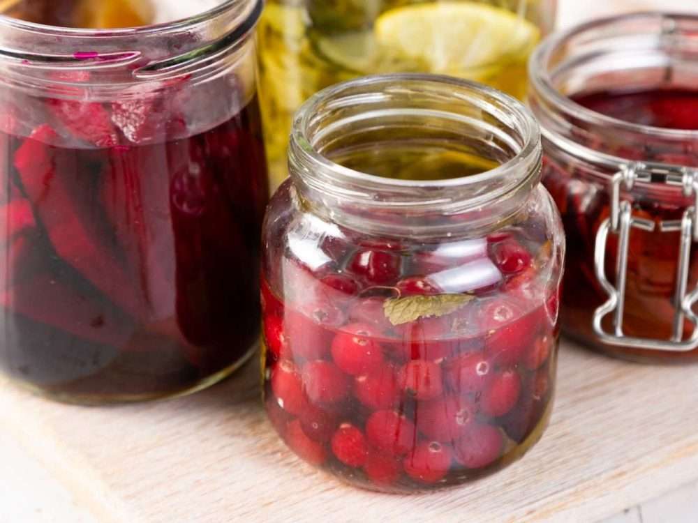 Jars of fruit steeping in syrup on a pale wooden board — cranberries, dark red fruit and a clamp-top jar.