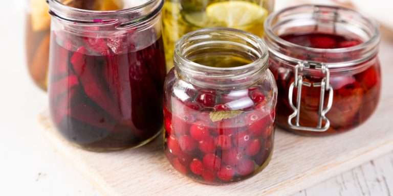 Jars of fruit steeping in syrup on a pale wooden board — cranberries, dark red fruit and a clamp-top jar.