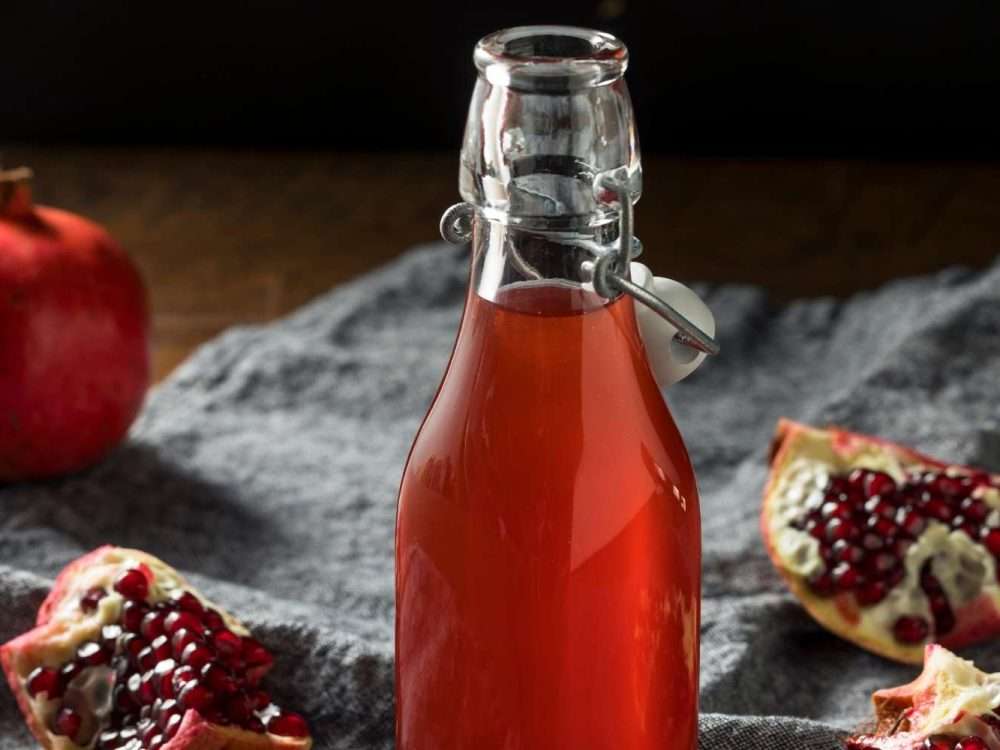 Homemade grenadine in a swing-top bottle on a grey cloth, with a whole pomegranate and cut wedges showing the seeds.