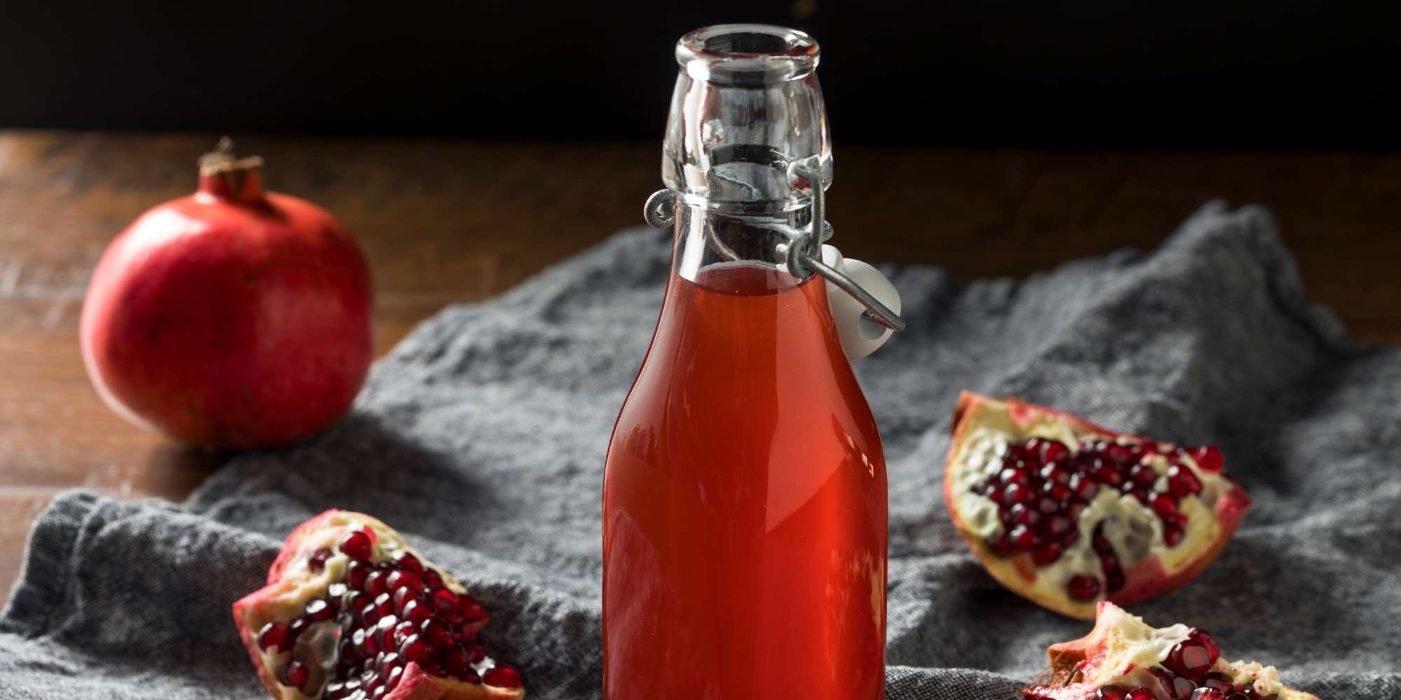 Homemade grenadine in a swing-top bottle on a grey cloth, with a whole pomegranate and cut wedges showing the seeds.