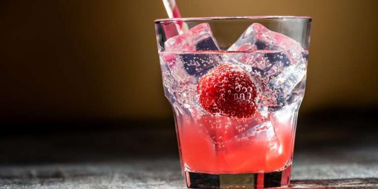 A Shirley Temple in a rocks glass — pale pink over ice cubes with a raspberry and a striped paper straw.
