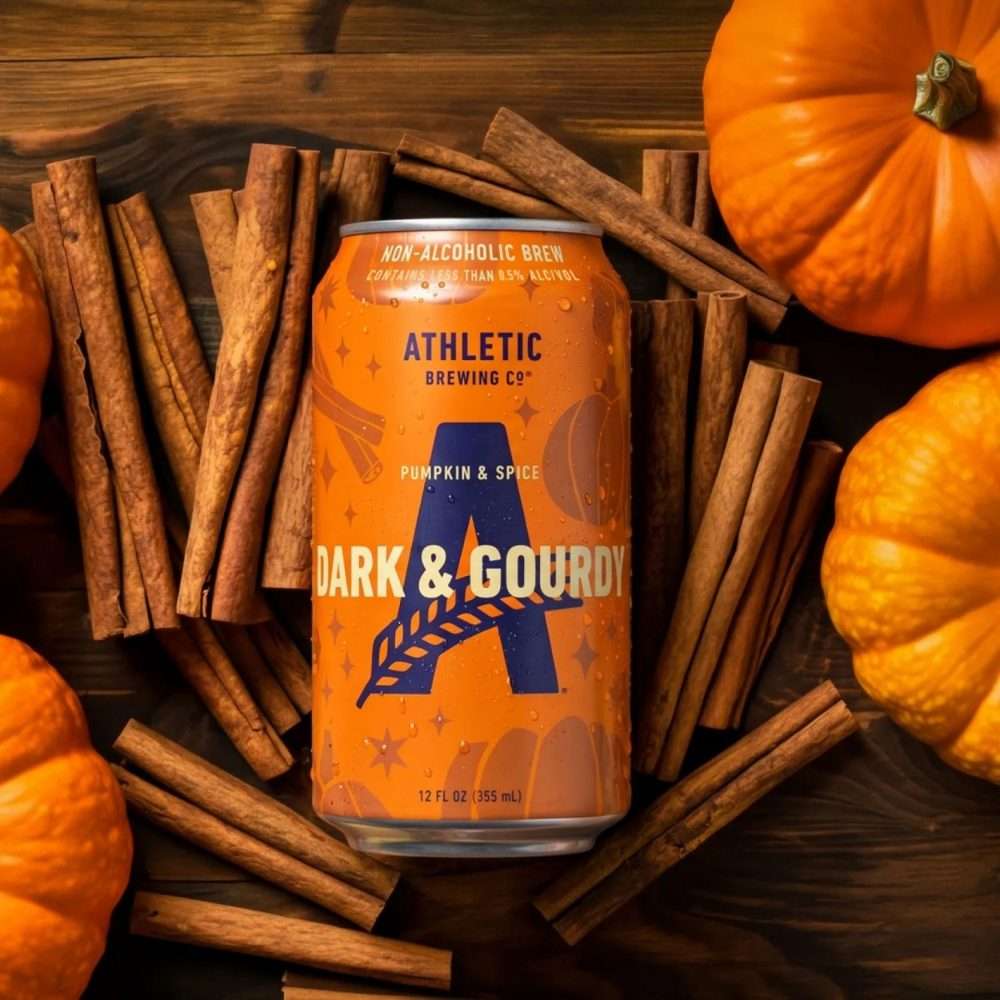 Can of Athletic Brewing Dark & Gourdy non-alcoholic brew lying on dark wood, framed by cinnamon sticks and small pumpkins