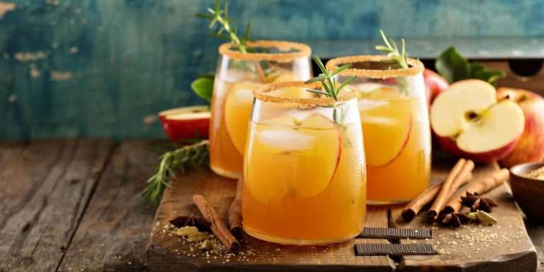 Three apple cider mocktails with cinnamon-sugar rims, apple slices and rosemary, beside apples and cinnamon sticks