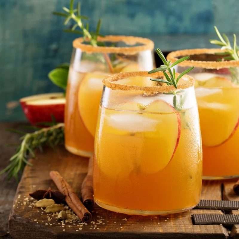 Three apple cider mocktails with cinnamon-sugar rims, apple slices and rosemary, beside apples and cinnamon sticks