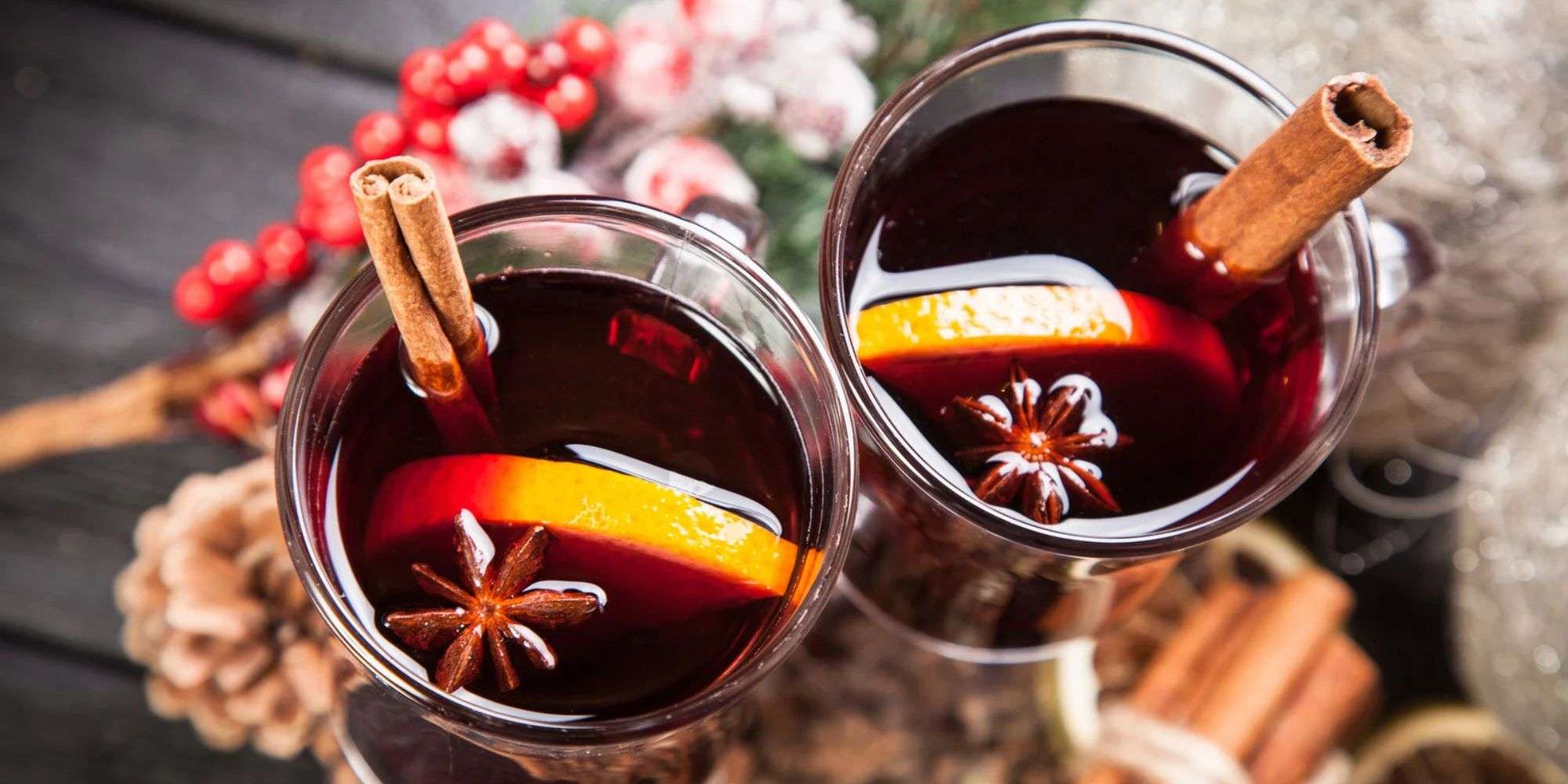 Two glass mugs of non-alcoholic mulled wine, each with an orange slice, star anise and a cinnamon stick, on a dark wooden table with red winter berries and pine cones.
