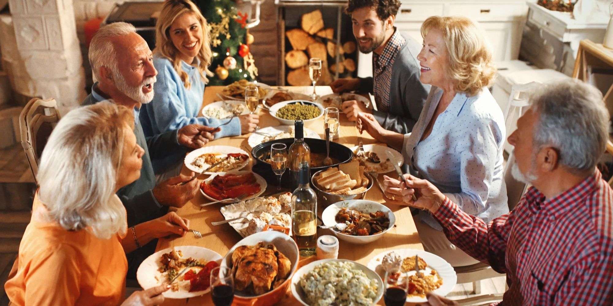 Family gathered around a holiday dinner table with a roast turkey, side dishes, bottles of white wine, and glasses of sparkling and red wine